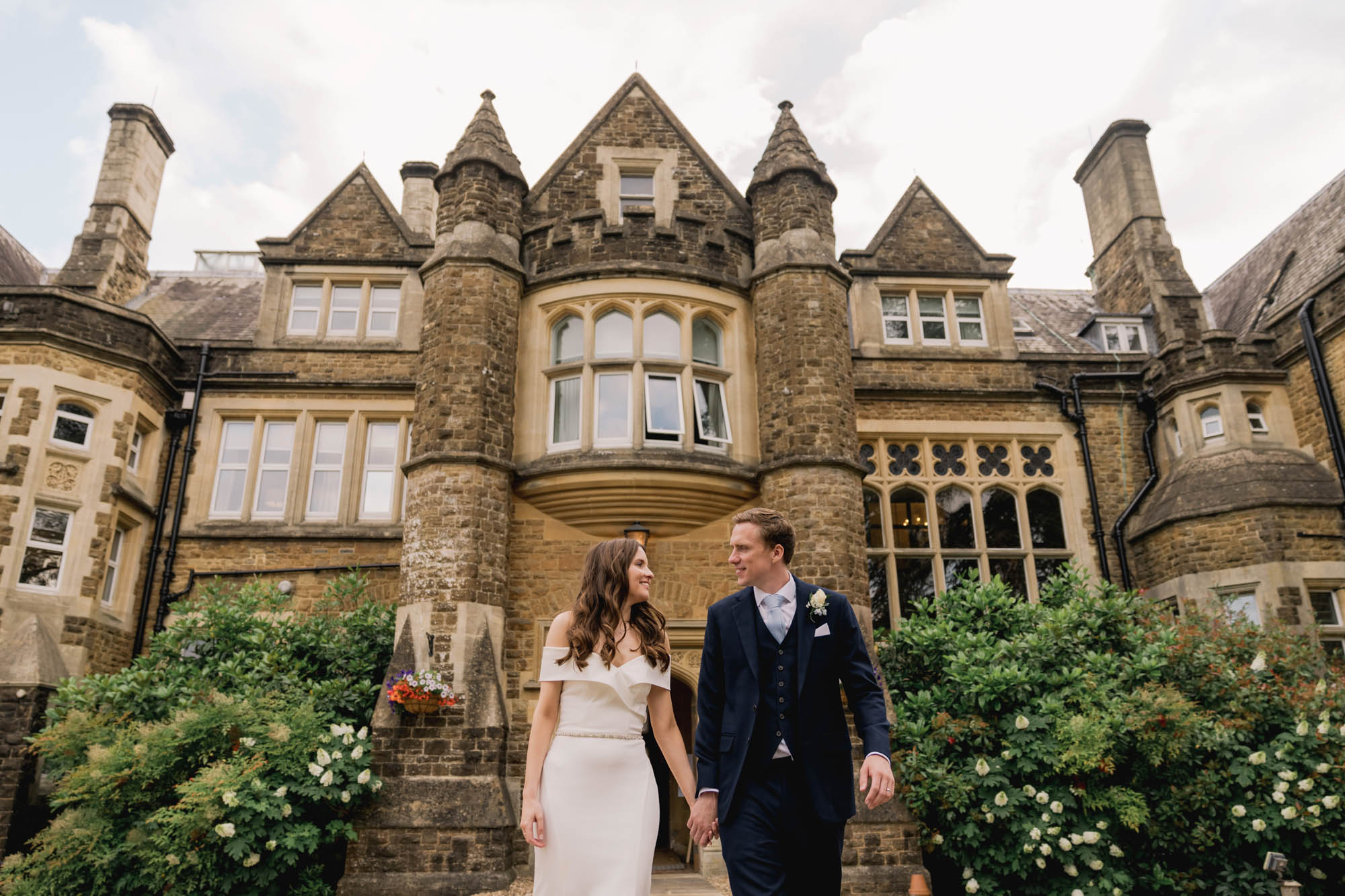 Bride and groom smiling whilst they take a stroll on their wedding day with Hartsfield manor in the background.