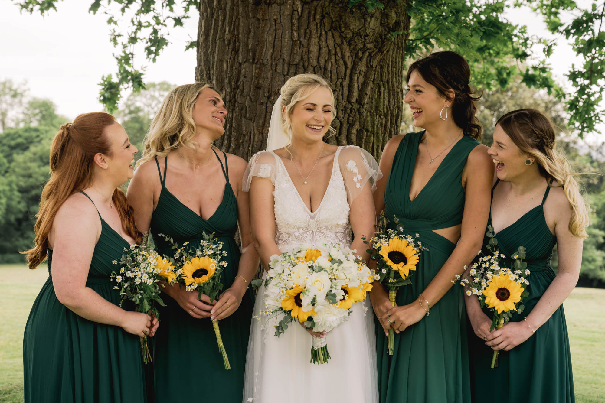 Bride laughs with her bridesmaids under the oak tree at Hartsfield manor in Betchworth.