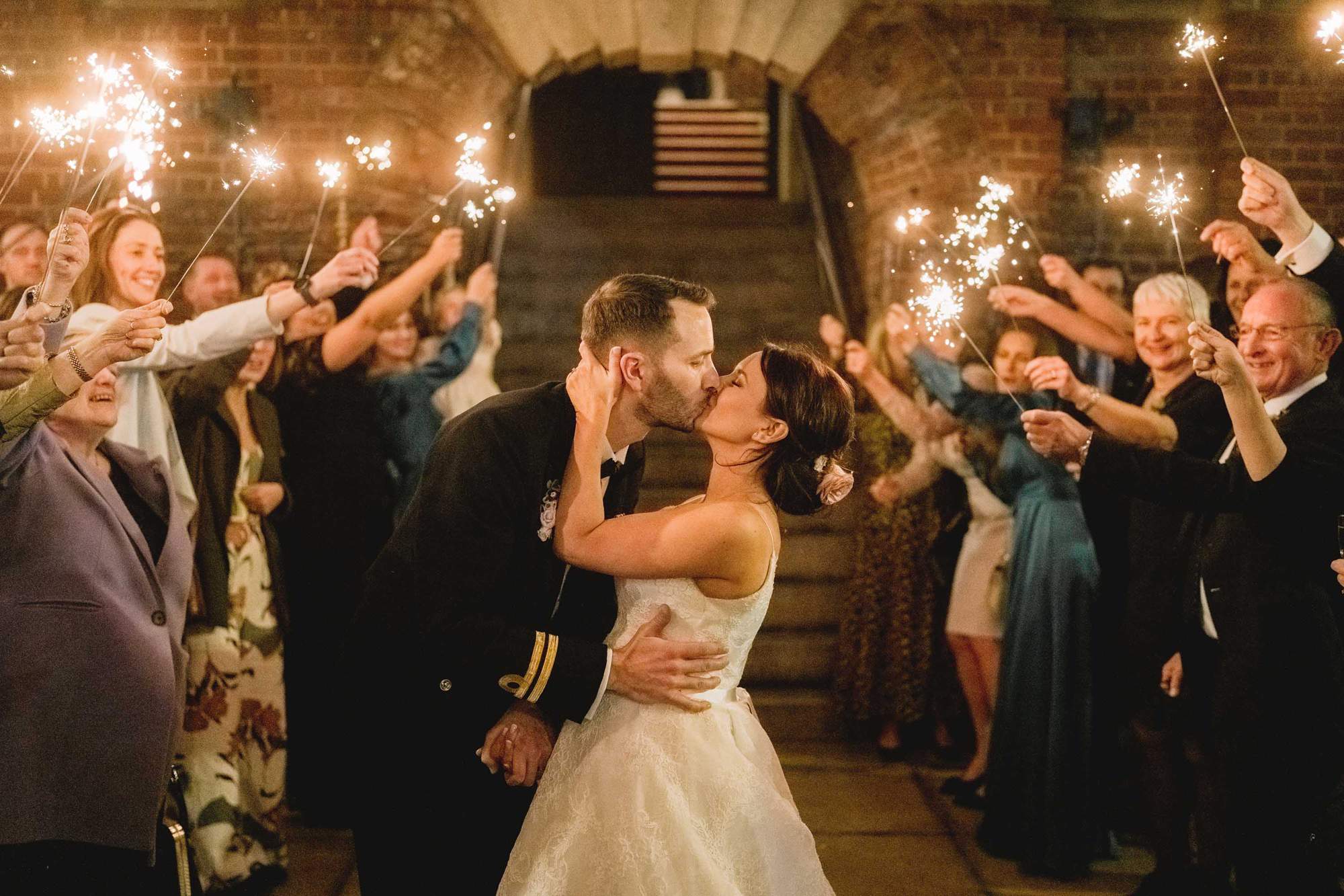Bride and groom kiss as their wedding guests cheer whilst holding sparklers.