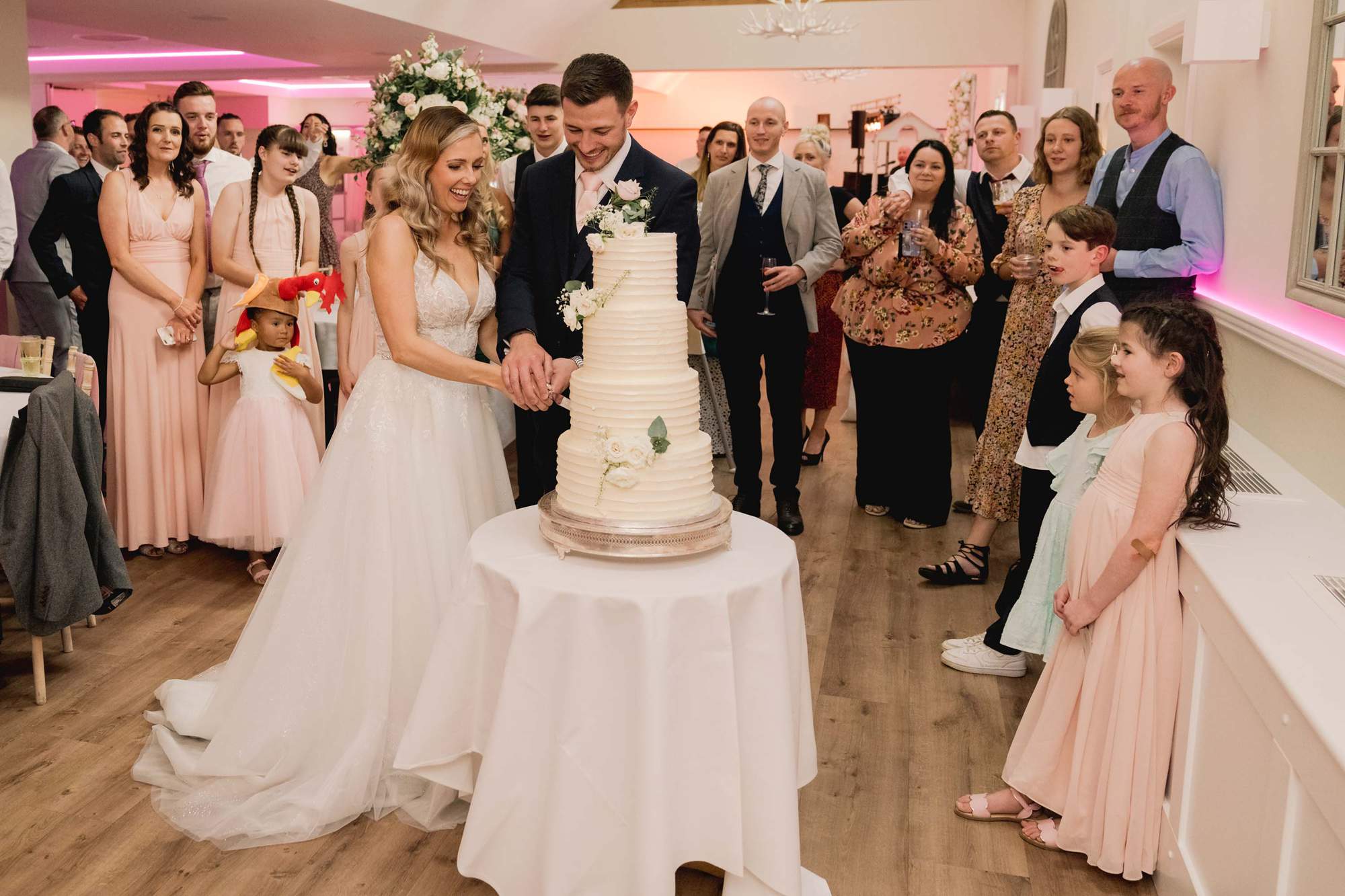 Bride and groom cut their wedding cake at Cottesmore Golf Club as their guests watch and cheer.