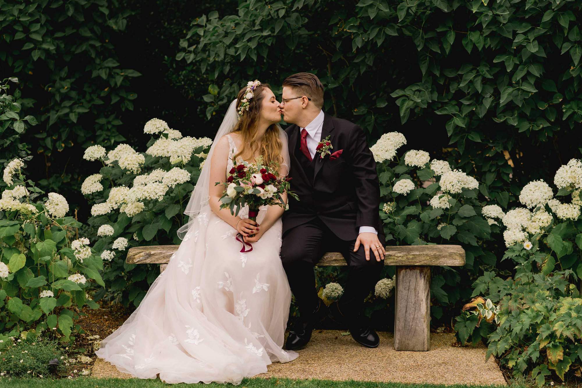 Bride and groom have a kiss on a bench on their wedding day at the Clock Barn venue in Hampshire.